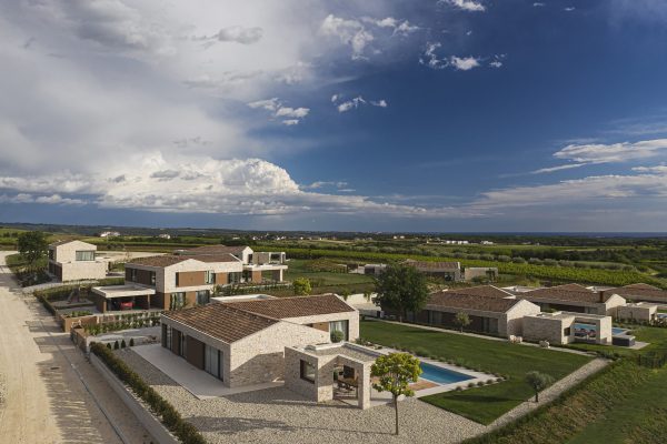 Zračni prikaz stambenog naselja s modernim kućama, krovovima prekrivenim crijepom, uređenim dvorištima, bazenom i otvorenim seoskim krajolikom pod djelomično oblačnim nebom.