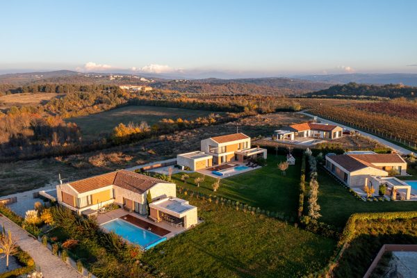 Zračni prikaz modernih kuća s bazenima, okruženih poljima i drvećem u ruralnom području pri zalasku sunca.
