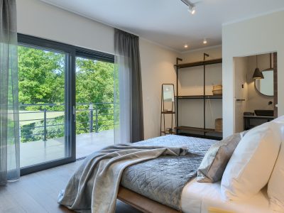 Moderna spavaća soba s velikim staklenim vratima koja vode na balkon, uredno poslanim krevetom, otvorenim ormarom i susjednom kupaonicom vidljivom u pozadini.
