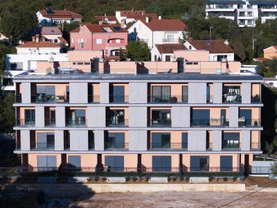 Moderna četverokatna stambena zgrada s balkonima, smještena naspram pozadine s kućama i zelenilom na padini.