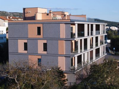 Moderna trospratna stambena zgrada s balkonima i sivom te breskvastom vanjštinom, okružena drvećem i smještena naspram brdovitog krajolika.