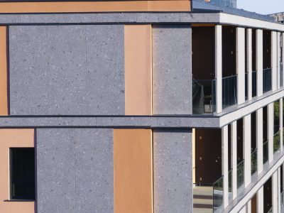 Moderna stambena zgrada sa sivim i narančastim vanjskim zidovima, staklenim balkonima i više katova vidljivih na jarkom dnevnom svjetlu.