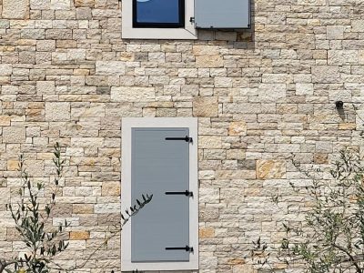 Kamena fasada zgrade s dvoja zatvorena siva kapka; lijevi kapak gornjeg prozora je otvoren, otkrivajući dio stakla. Ispred zida rastu neke biljke.