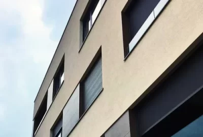 Moderna bež zgrada s velikim pravokutnim prozorima i tamnim okvirima prozora, prikazana iz niskog kuta na pozadini oblačnog neba.