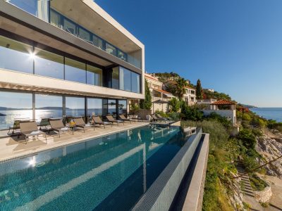 Moderna vila uz more s velikim prozorima i beskonačnim bazenom s pogledom na ocean, okružena ležaljkama i obližnjim obalnim kućama pod vedrim plavim nebom.