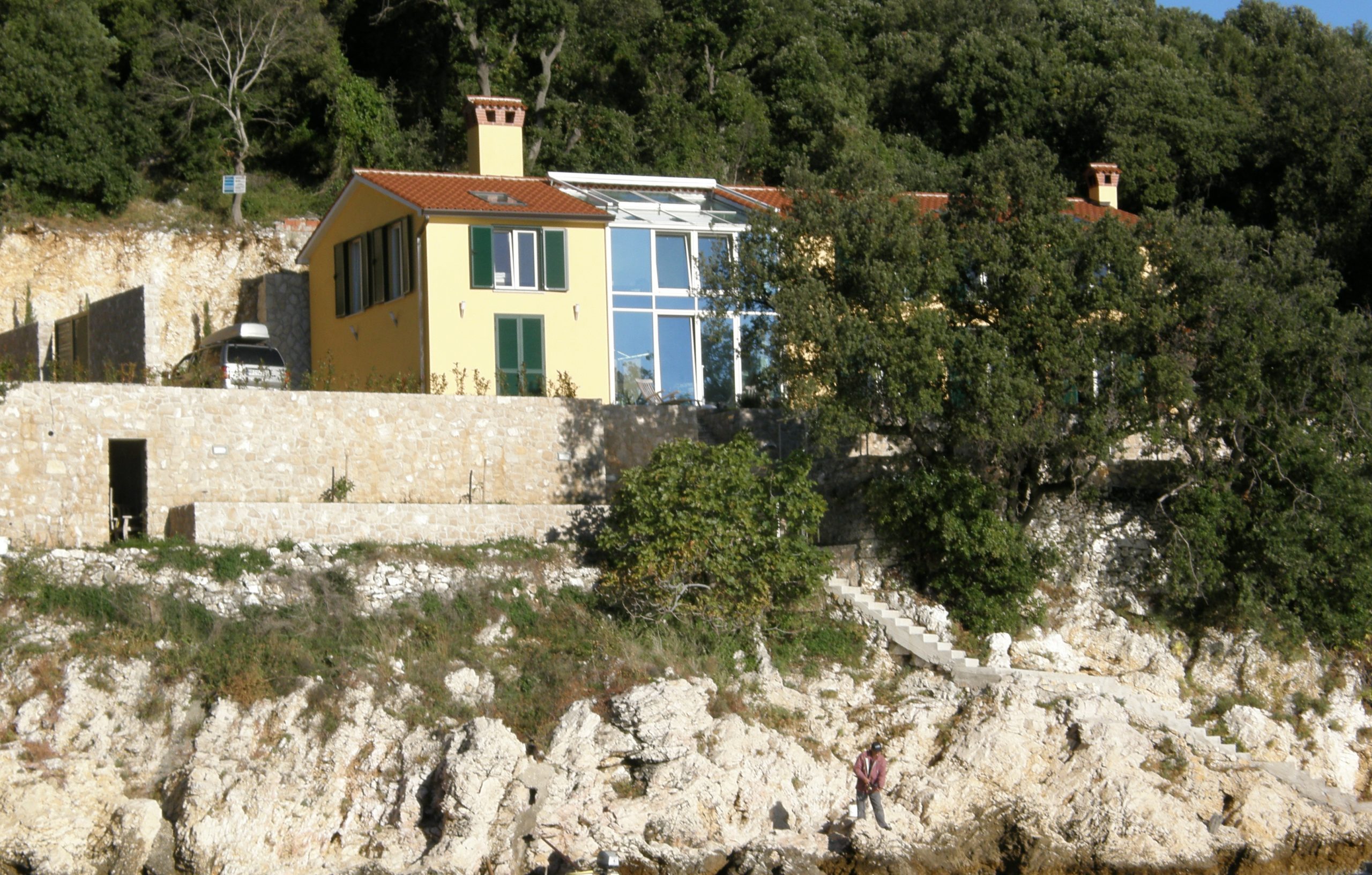 Žuta kuća s crvenim krovom i staklenim prozorima smještena je na stjenovitoj, drvećem obrasloj padini iznad vode, s malim čamcem privezanom ispod.