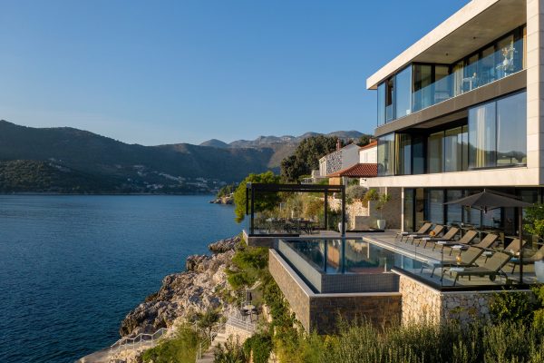 Moderna vila uz obalu s balkonima od stakla, beskonačnim bazenom, ležaljkama i panoramskim pogledom na more i udaljene planine pod vedrim plavim nebom.