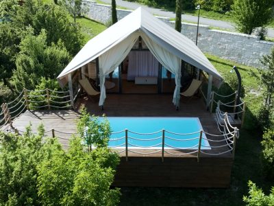 Glamping šator s otvorenim zavjesama stoji na drvenoj terasi, uz pravokutni bazen, okružen zelenilom i ogradom od užeta.