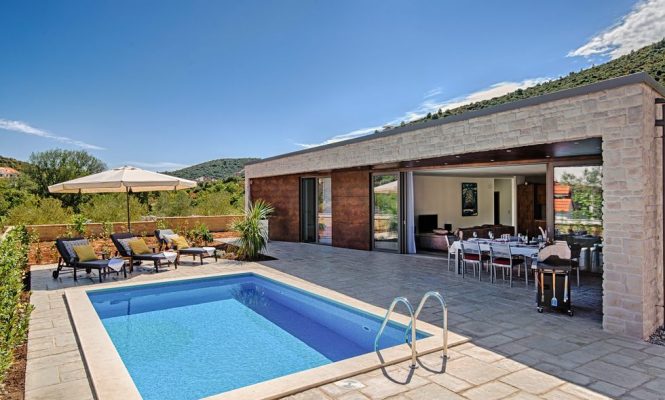 Moderna vila s malim pravokutnim bazenom, ležaljkama, vanjskim blagovaonskim prostorom i kliznim staklenim vratima, smještena naspram zelenih brežuljaka pod vedrim plavim nebom.