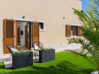 Mali popločani patio s dvije crne pletene stolice i stolom nalazi se na umjetnoj travi ispred bež kuće s drvenim kapcima i otvorenim vratima koja vode unutra. U prvom planu nalazi se palma u tegli.