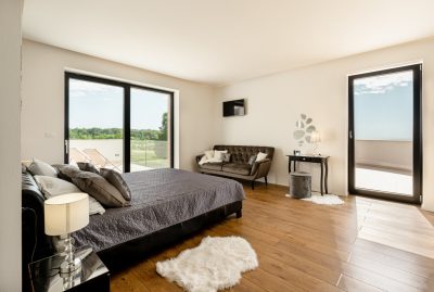 Moderna spavaća soba s velikim krevetom, sivim prekrivačem, malom sofom i kliznim staklenim vratima koja se otvaraju prema vanjskom pogledu; drveni podovi i minimalistički dekor.